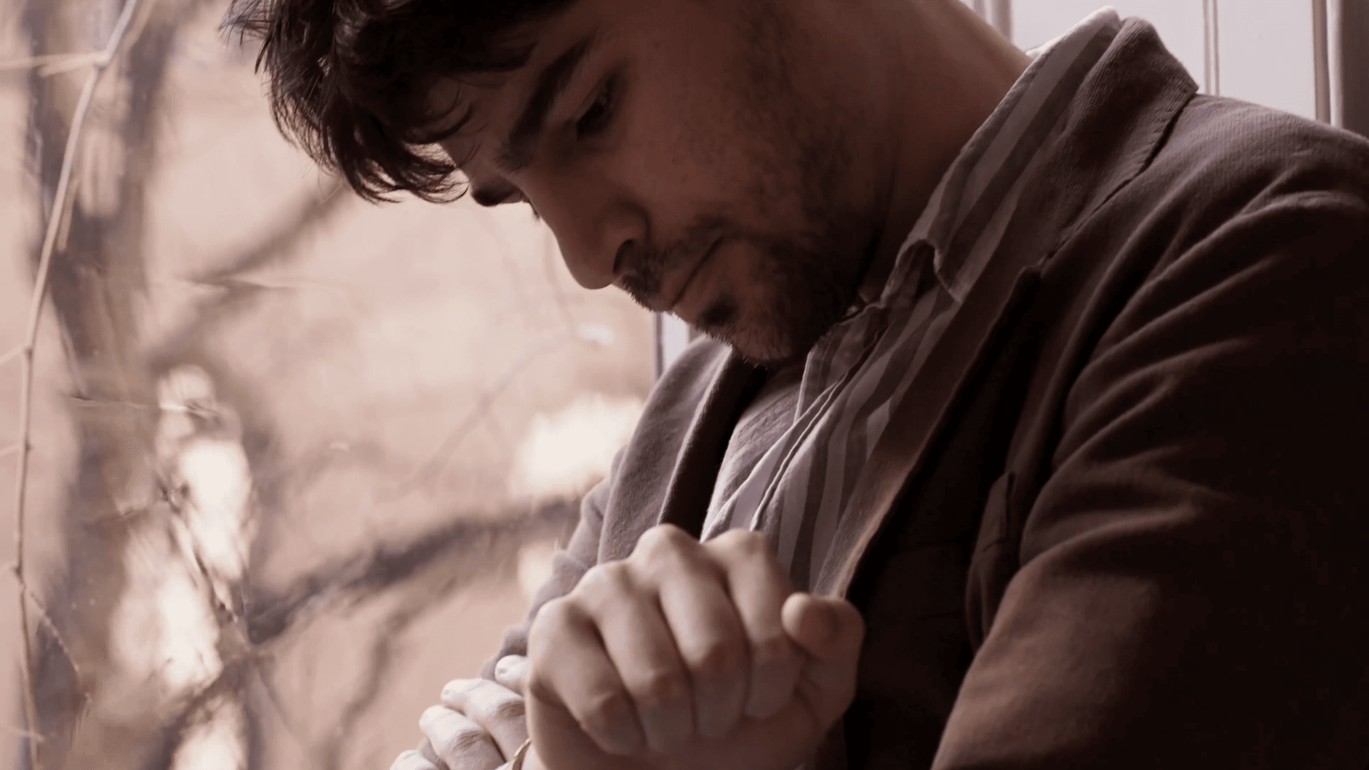 man wearing a brown jacket looking down at watch in front of a sunny window