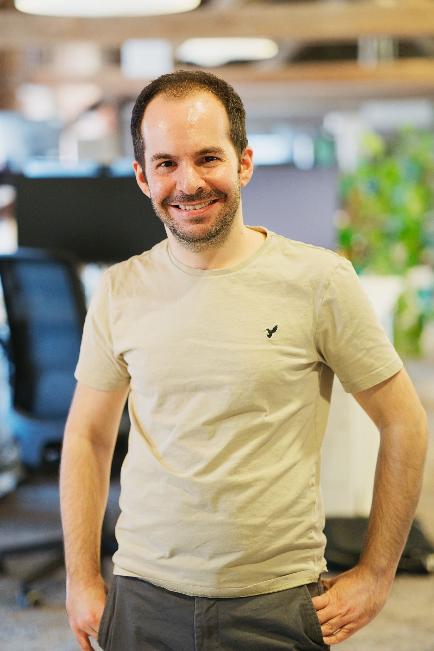 Ein Mann mit einem hellbeigen T-Shirt steht in einer Büroumgebung und hat die Hände in die Hüften gestemmt.
