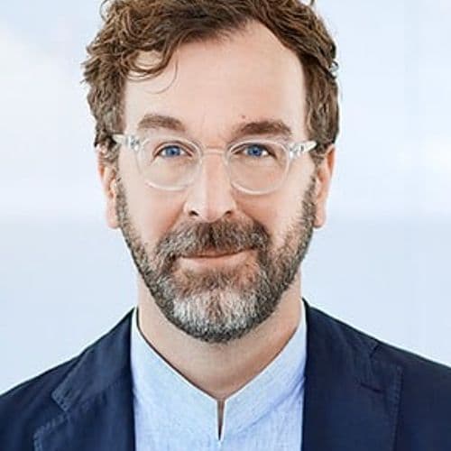 Man in a suit jacket and light shirt standing in front of a plain background.