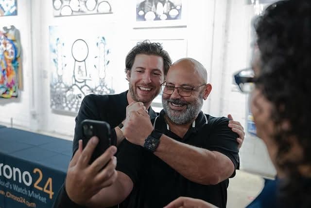 two men in black shirts at a conference posing  in front of a bright wall showing off their watches and taking a selfie