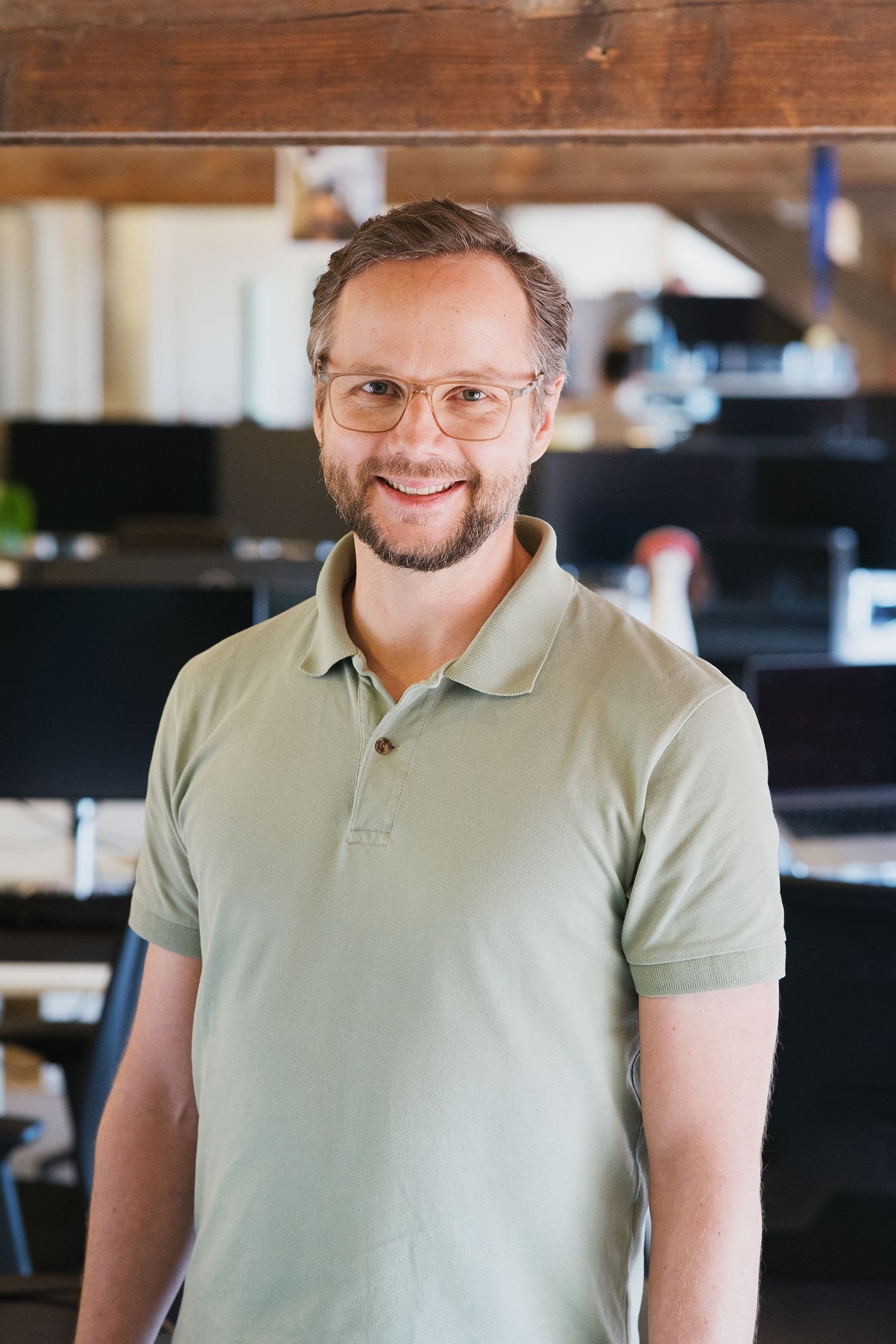Ein Mann mit hellgrünem Polohemd steht in einem Büro mit sichtbaren Holzbalken und Computerbildschirmen im Hintergrund.