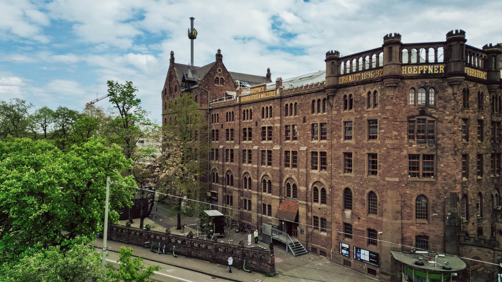 Ein großes historisches Backsteingebäude mit vielen Rundbogenfenstern und Türmen, umgeben von grünen Bäumen. An der Fassade stehen die Schilder „Hoepfner“ und „Bierbrauerei & Mälzerei“.