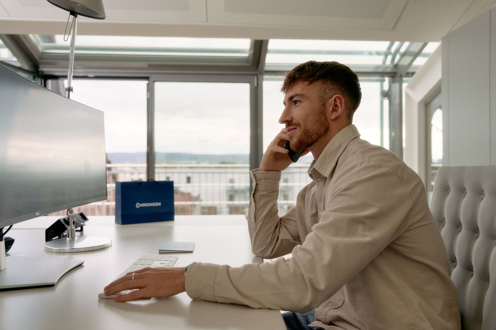 man wearing a tan shirt sat at a computer and holding a smartphone to his ear