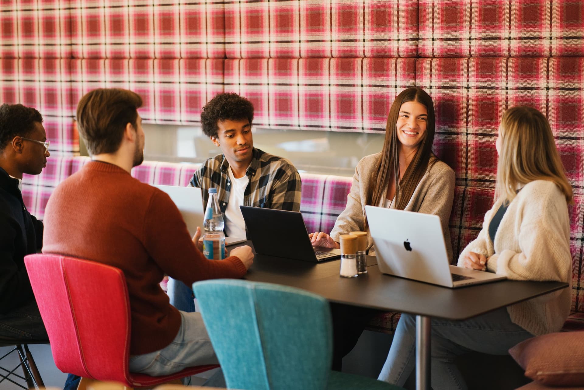 Eine Gruppe von vier Personen sitzt zusammen an einem Tisch in einer modernen Bürolounge und unterhält sich. Auf dem Tisch stehen Laptops, Wasserflaschen und Kaffeetassen, die Wand dahinter hat ein rot kariertes Muster.
