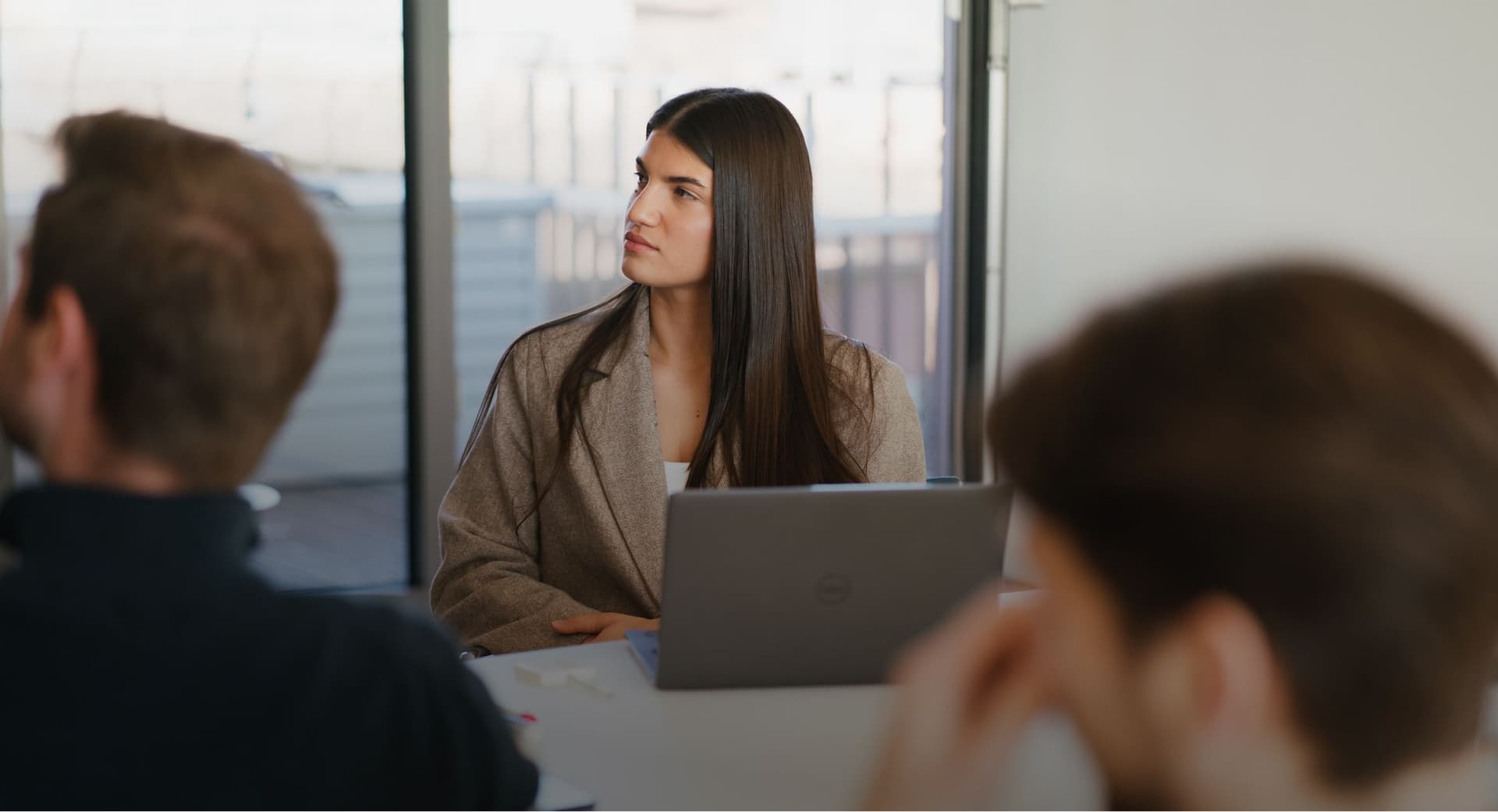 Frau mit langen Haaren im braunen Blazer sitzt bei einer Besprechung mit Laptop am Tisch.