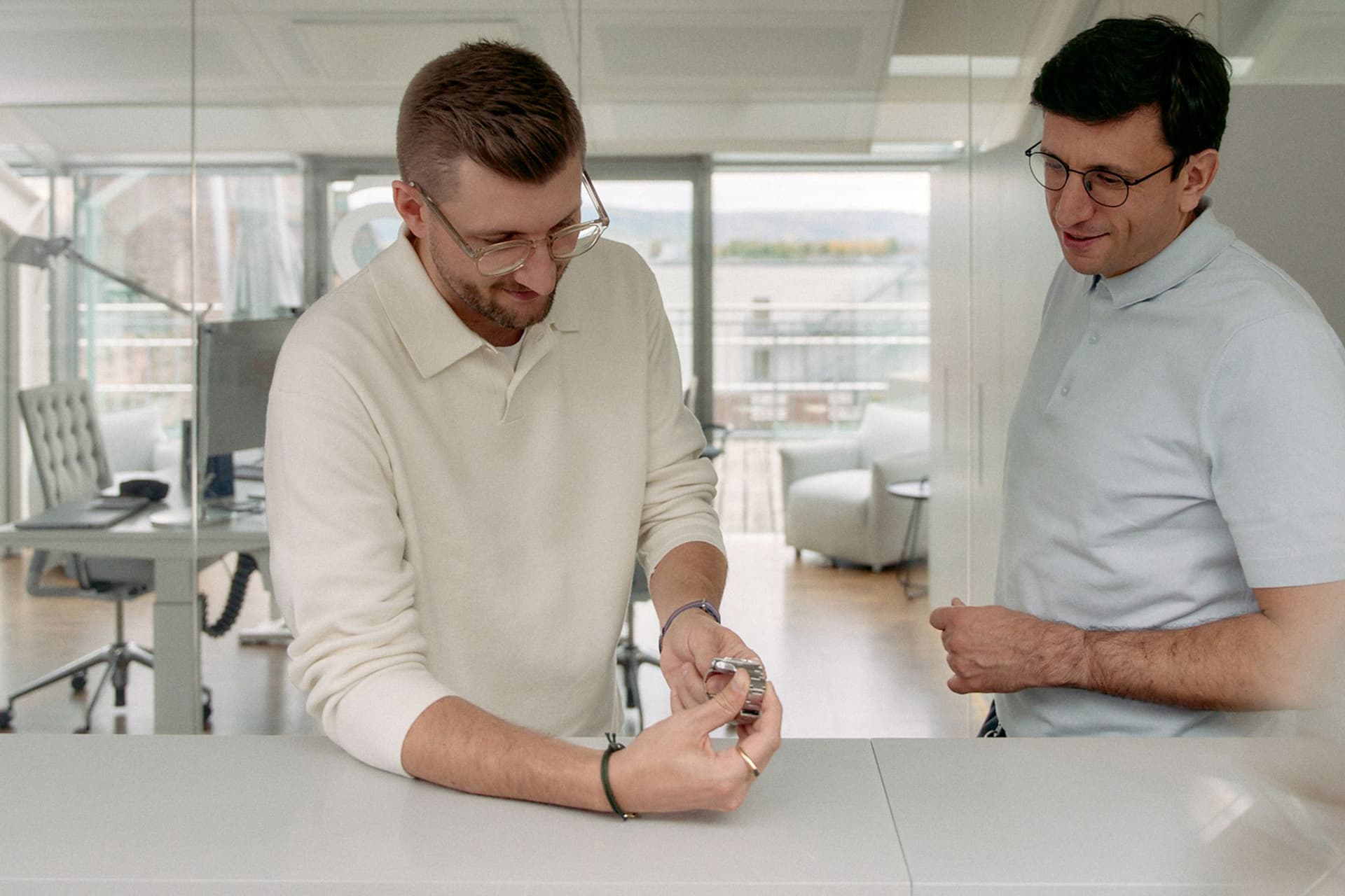two men wearing light shirts examining a watch in a bright room with glass windows