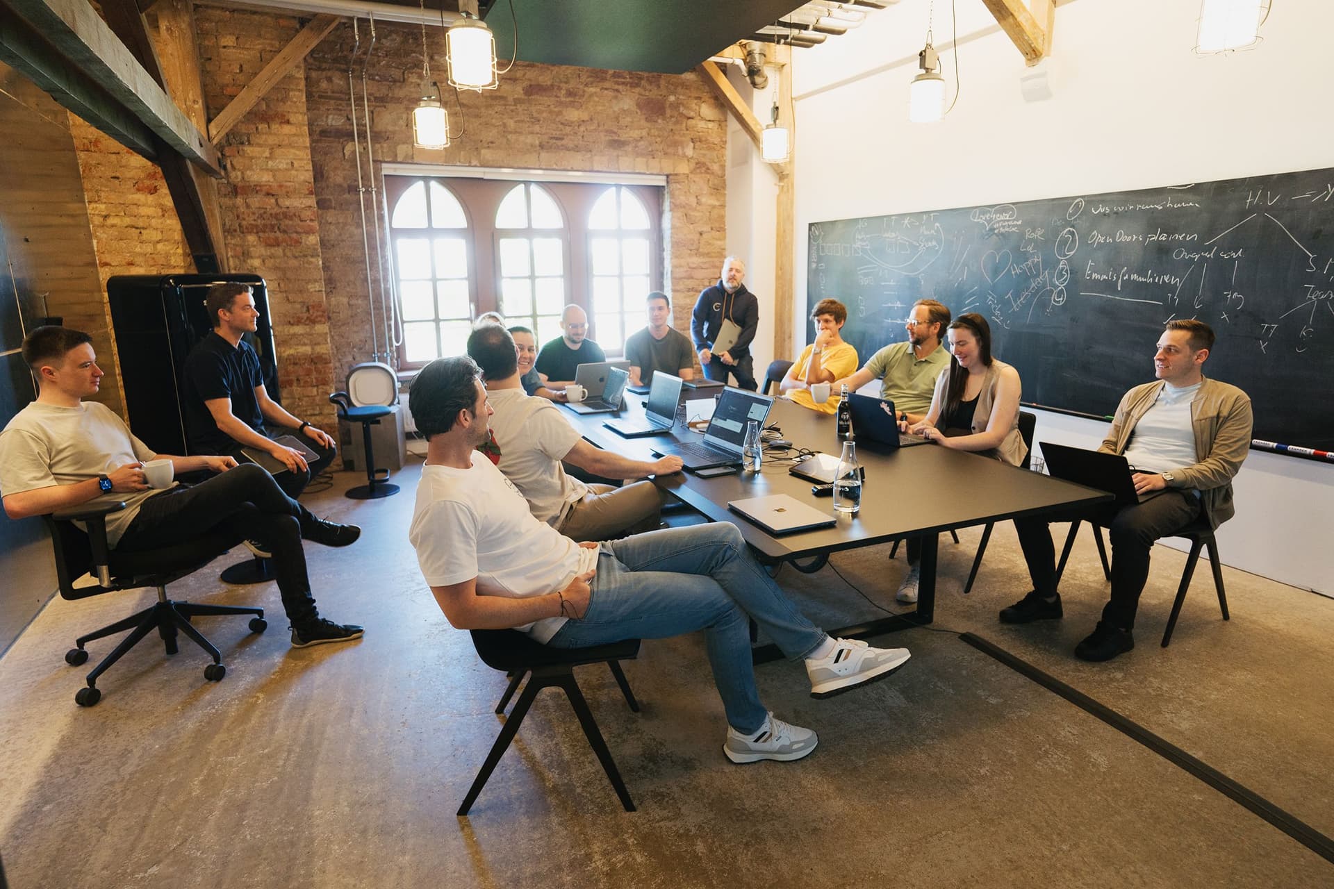 Several people are having a meeting around a large table in a modern room with brick walls, wooden beams, and a chalkboard.
