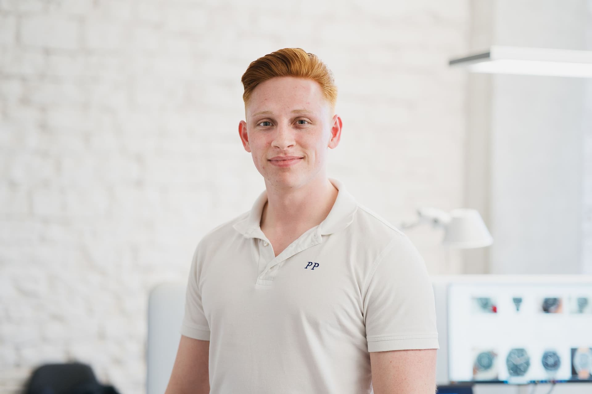 Ein Mann mit kurzen roten Haaren trägt ein weißes Polohemd mit den Initialen „PP“ und steht in einem hellen Büro mit weißer Backsteinwand.