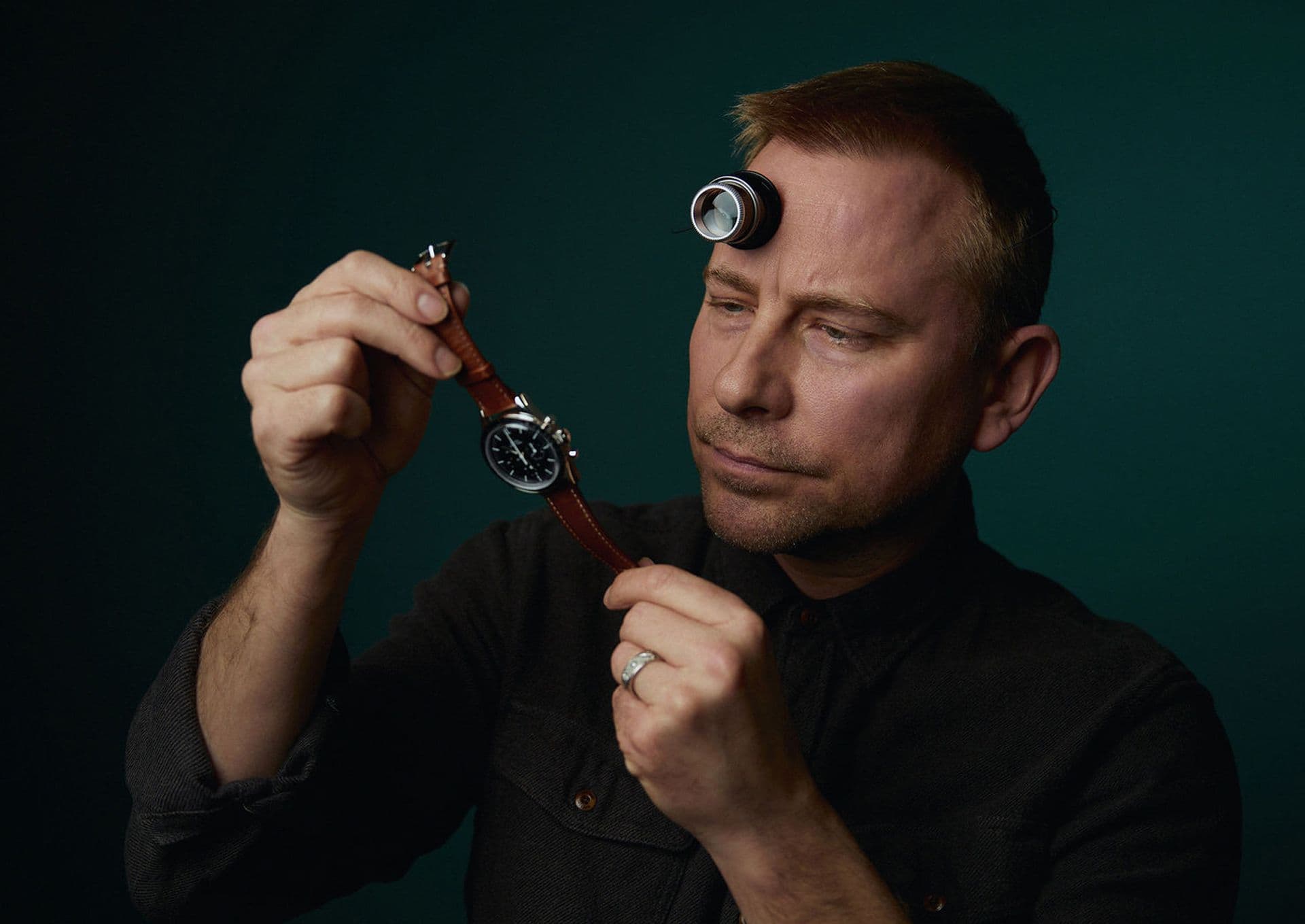 man wearing dark shirt and loupe holding up a leather strapped watch against a dark green background