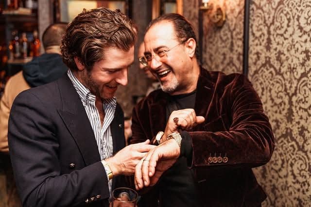 men wearing dress attire smiling at each other while showing off a watch