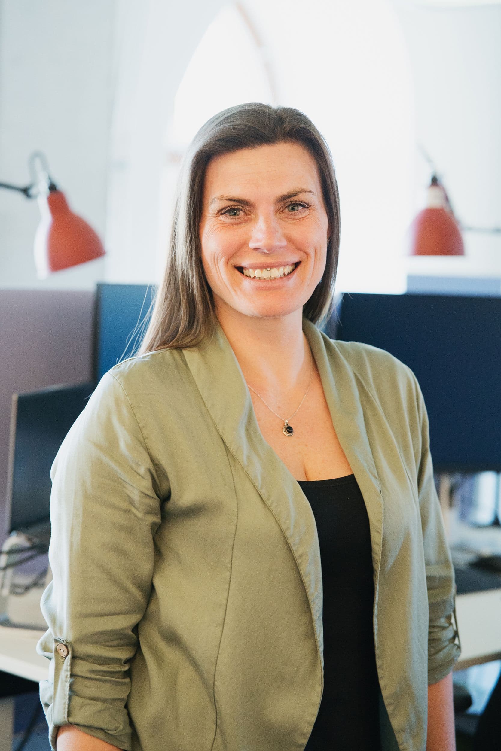 Eine Frau mit langen braunen Haaren, die einen hellgrünen Blazer über einem schwarzen Top trägt, steht in einem Büro mit Schreibtischen und roten Lampen im Hintergrund.