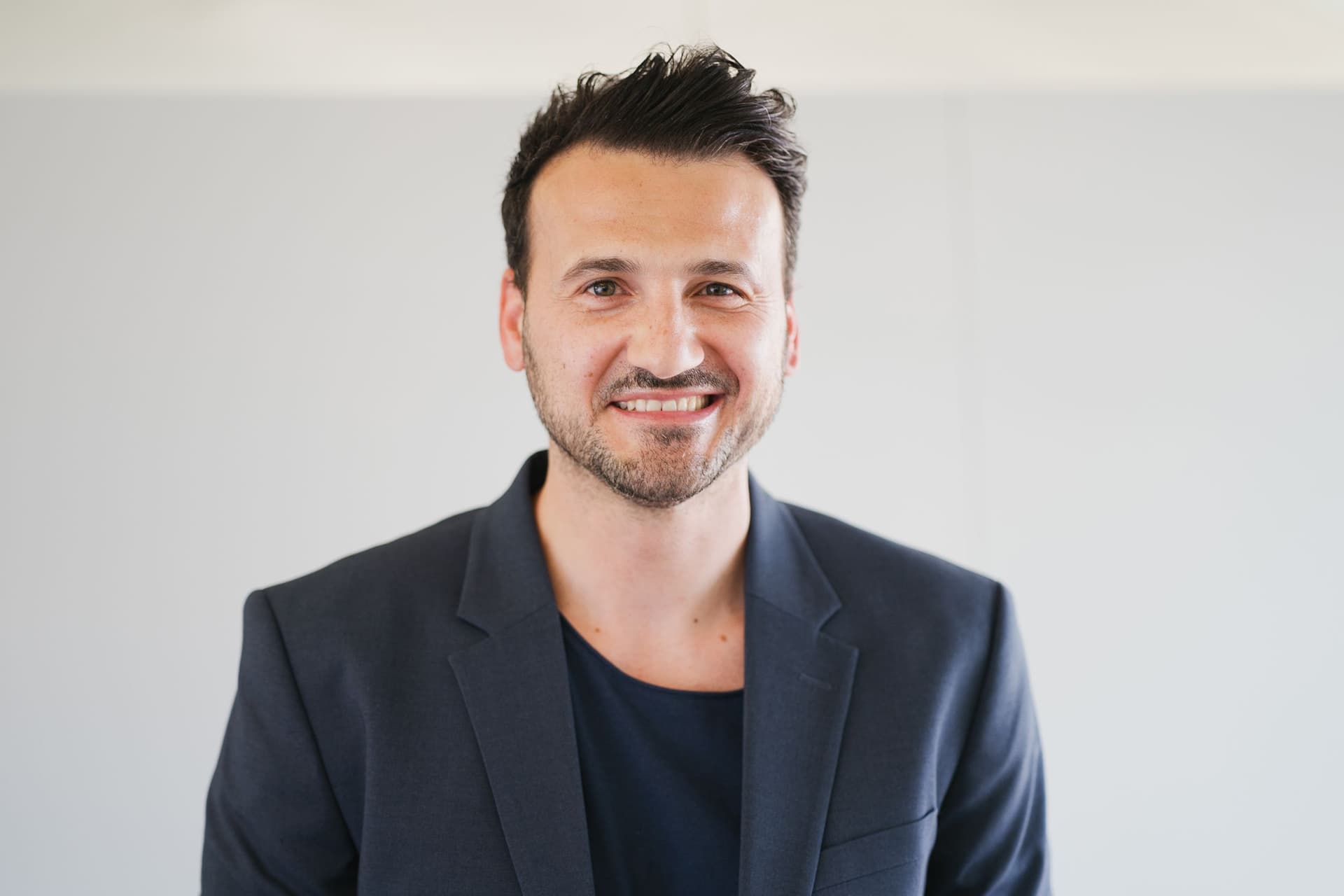 A man with dark hair wearing a dark blazer and a black shirt stands in front of a plain light background.