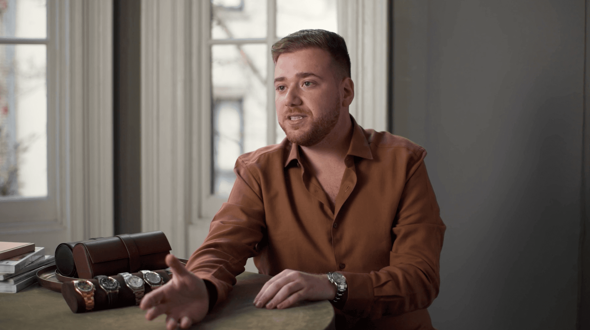 man in a brown shirt sat at a table filled with watches with a bright window behind