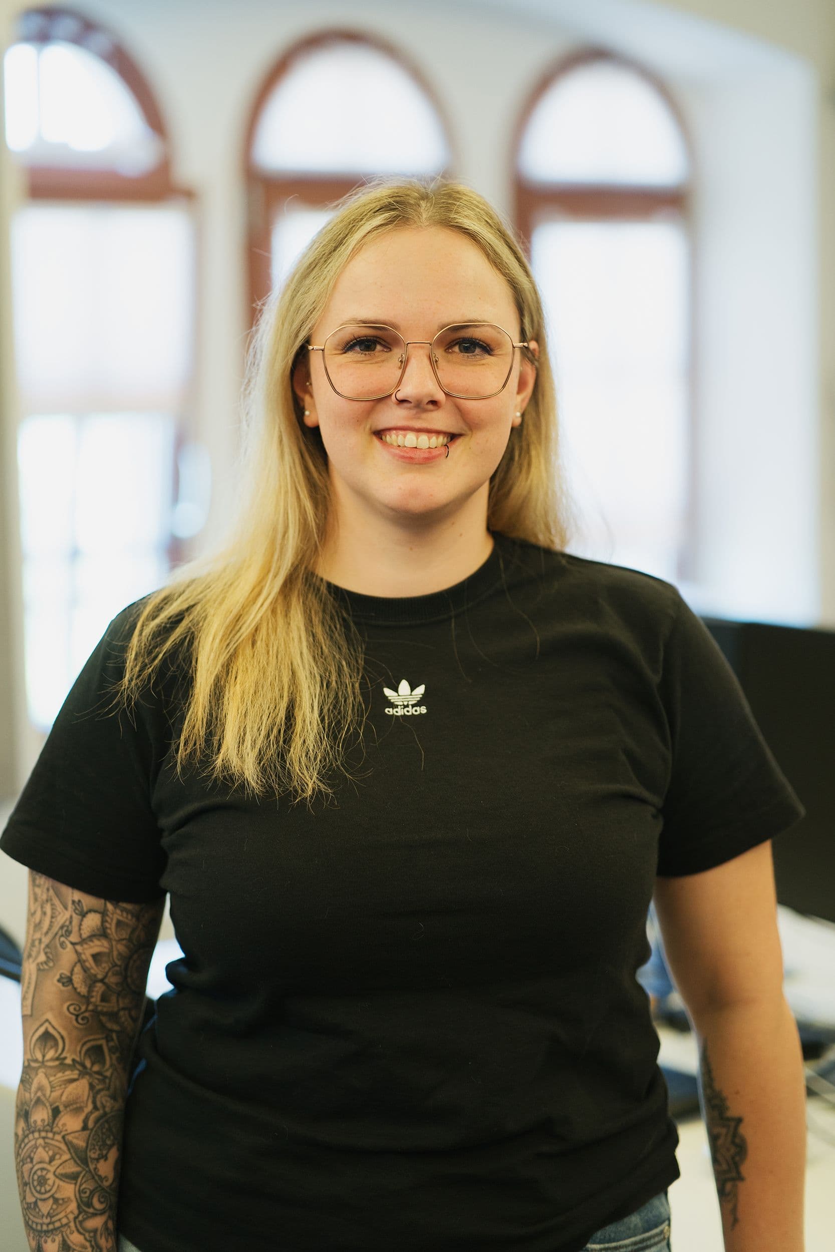 Eine Frau mit langen blonden Haaren und einem schwarzen Adidas-T-Shirt steht vor hellen Fenstern. Sie hat ein Tattoo am linken Arm.