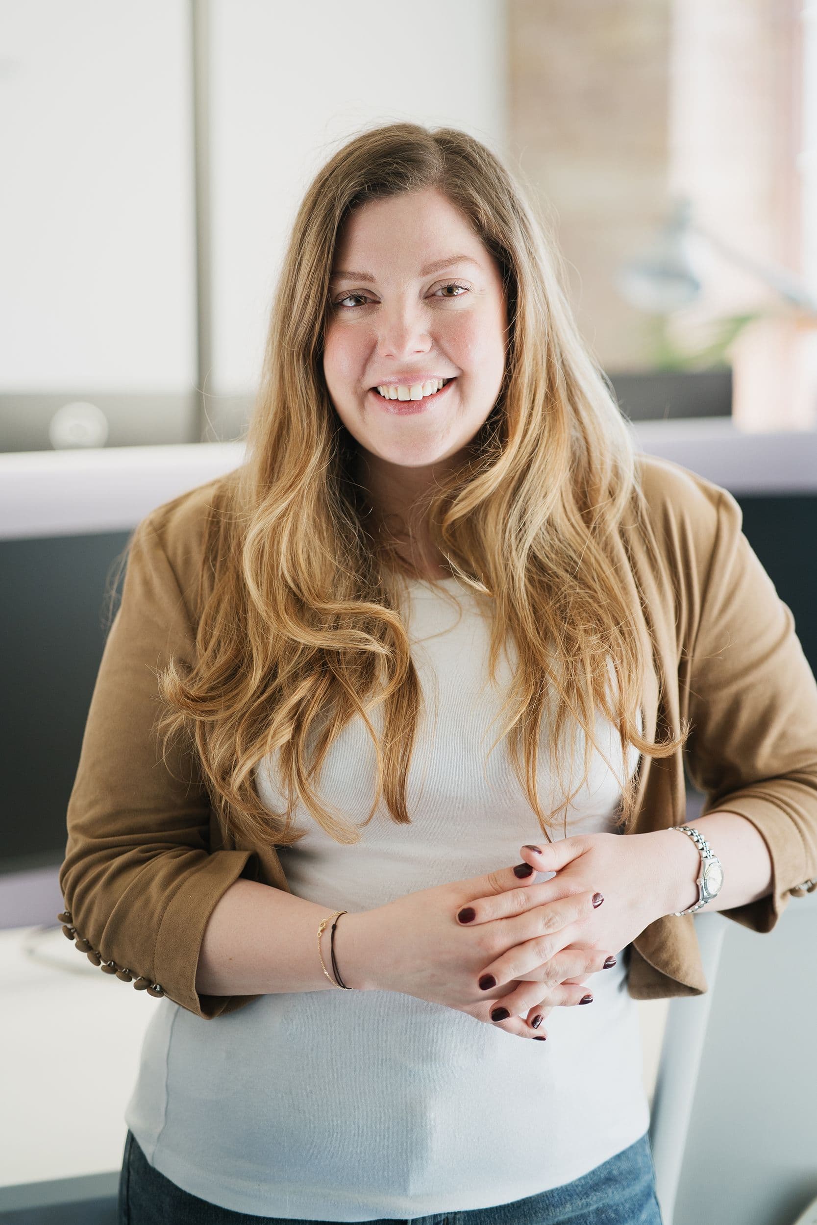 Eine Frau mit langen, welligen blonden Haaren trägt einen hellbraunen Blazer und ein weißes Oberteil und steht mit gefalteten Händen im Büro.