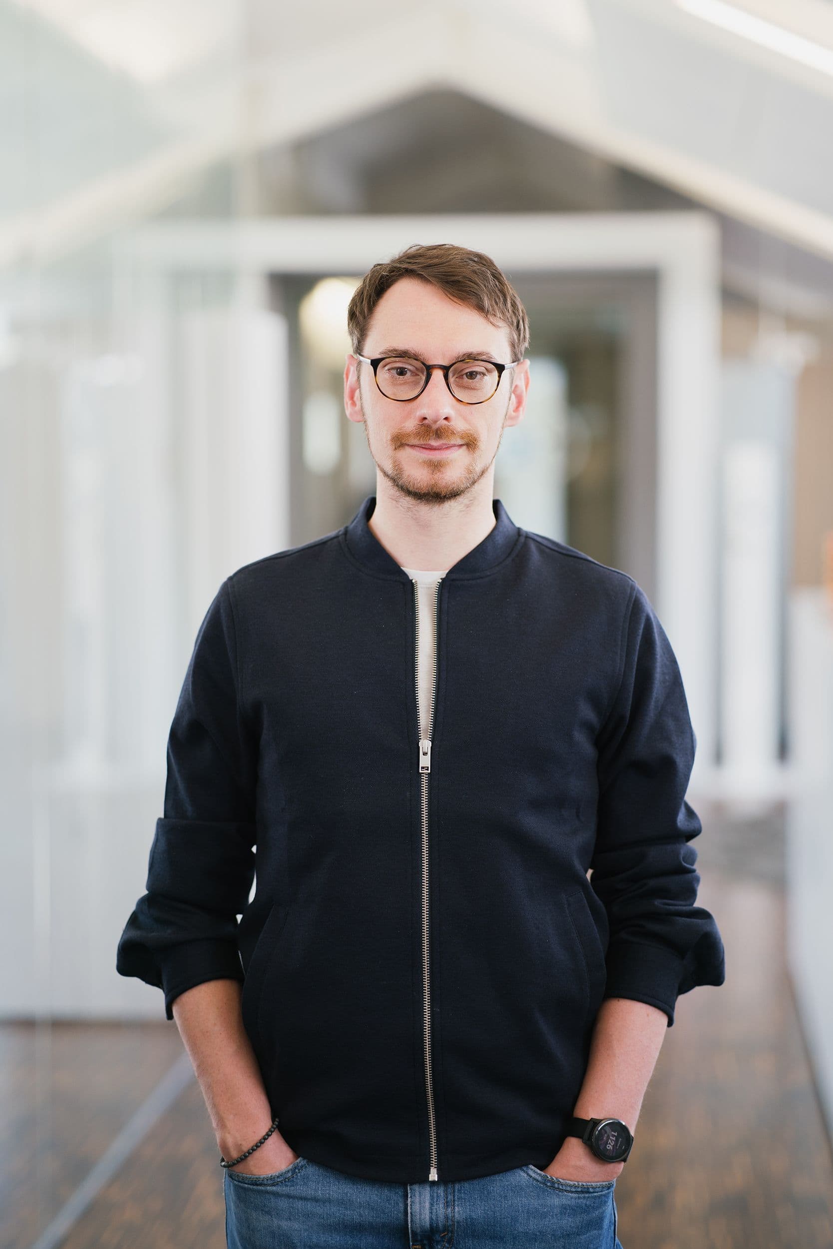 Ein Mann mit dunkelblauer Jacke mit Reißverschluss und Jeans steht mit den Händen in den Taschen in einem modernen Flur.