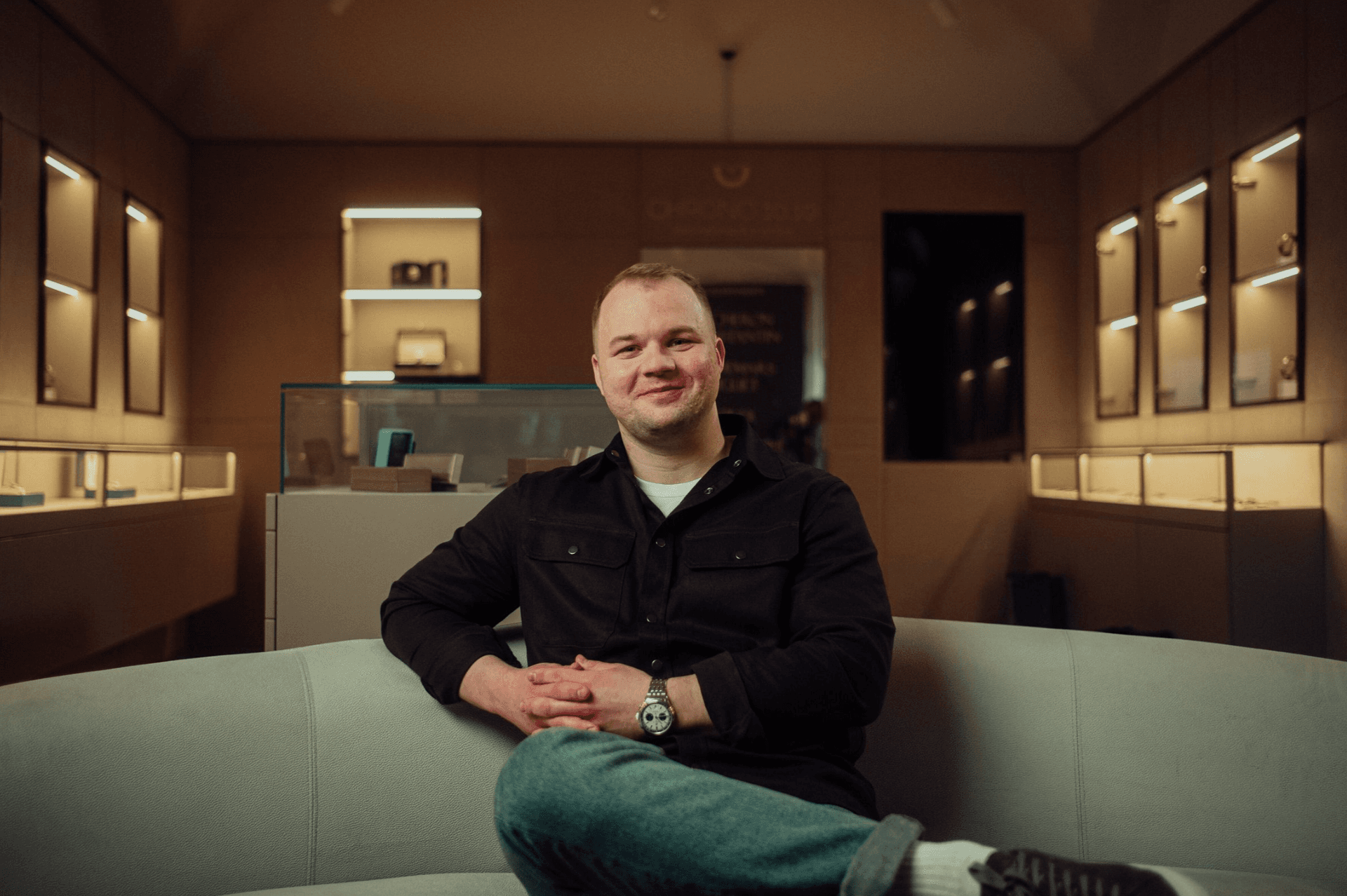 man in a dark button up shirt and jeans sat cross legged on a light colored couch in a dark showroom